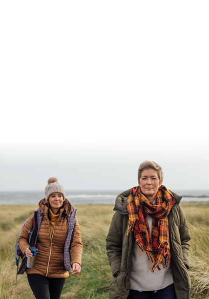 Two mature women wearing warm, casual, outdoor clothing and accessories on a day out in Northumberland. They smile and enjoy the view as they walk through the British countryside. One of them has short blond hair as she is in recovery after beating breast cancer.