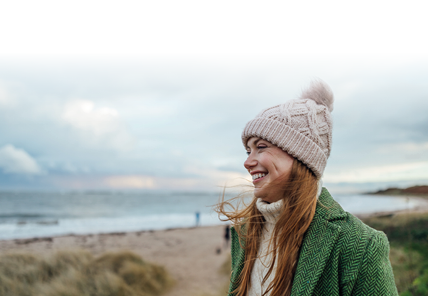 A side-view shot of a young female adult smiling whilst on a beach walk at Newton-by-the-Sea in Northumberland, North East England. She is looking straight ahead, to the left of the frame, with a wide contented smile. She is wearing a winter coat and woollen bobble hat, and behind her is a view of the beach and sea beyond.