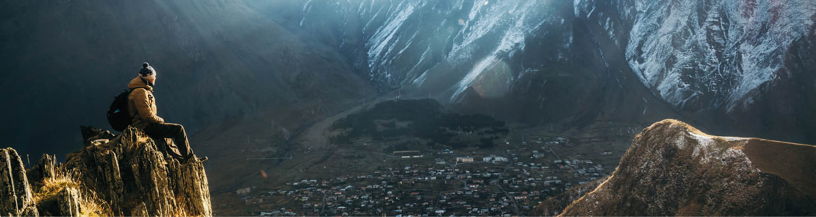 Young tourist in bright hat, black trousers with a backpack sit on cliff's edge and looking at the misty mountain village and glacier at sunrise, Stepantsminda, Georgia