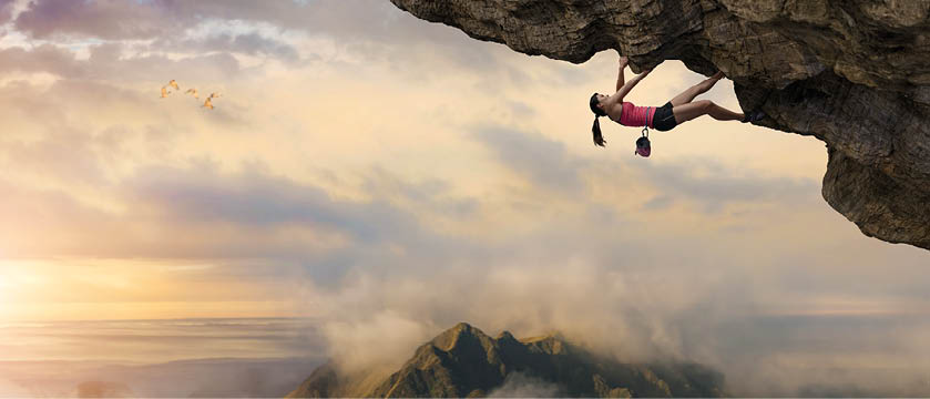 A woman free climber climbing the underside of an overhang of rock, high above a mountain peak. The free climber is wearing a sports vest and shorts, climbing shoes and is wearing a chalk bag, and is climbing as an extreme sport without safety equipment under a beautiful dawn sky.
