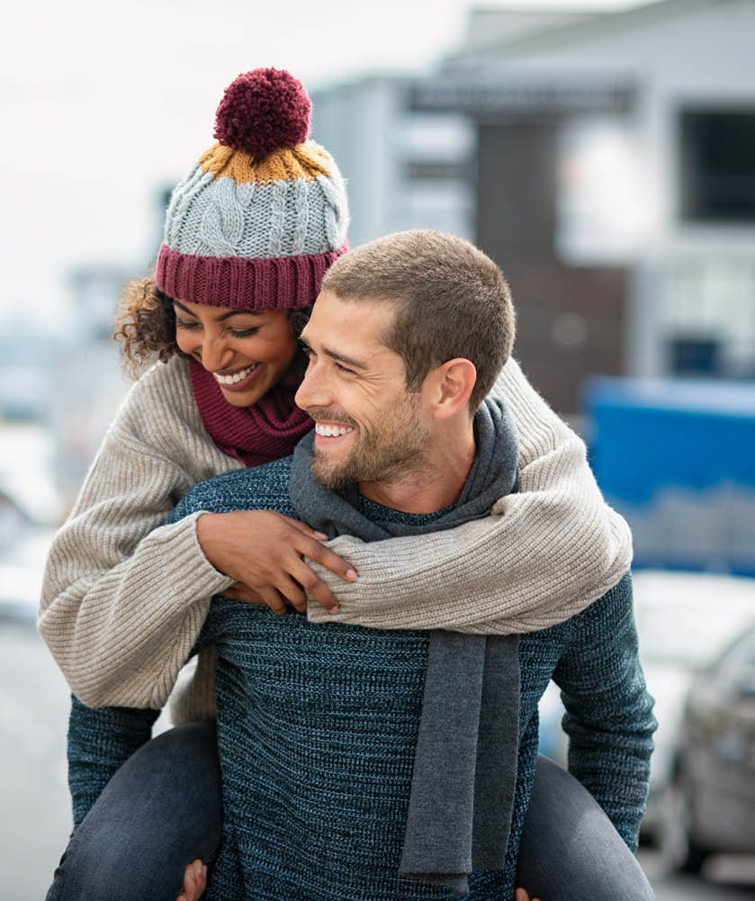 Smiling man giving piggyback ride to woman in the city  Young multiethnic couple in cold clothes walking in street and having fun  Cheerful girlfriend with wool cap and boyfriend in sweater enjoying winter together outdoor 