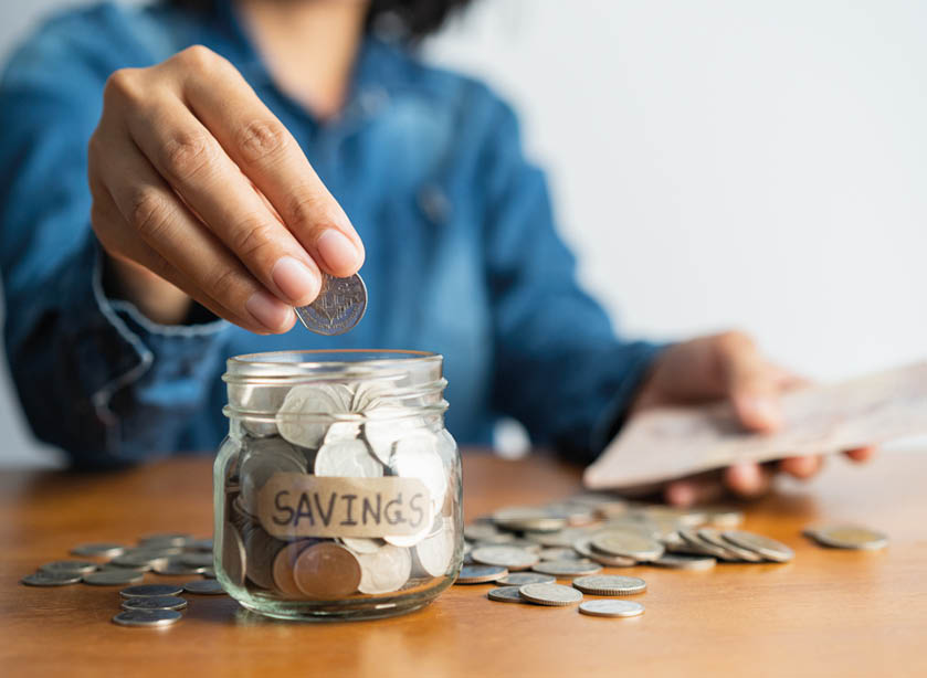 The woman hand is putting a coin in a glass  bottle and a pile of coins on a brown wooden table,Investment business, retirement, finance and saving money for future concept 