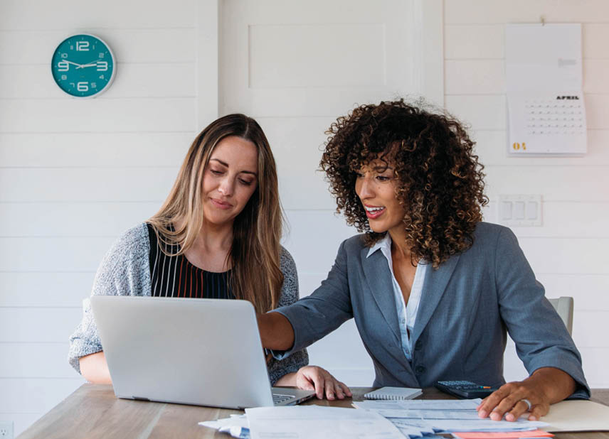 A female financial advisor sits with a woman at her dining room table with laptop and financial reports helping her monthly budget and investments  She is showing her client how to read a financial report as she works on her computer to do monthly finances, pay taxes and save money for the future 