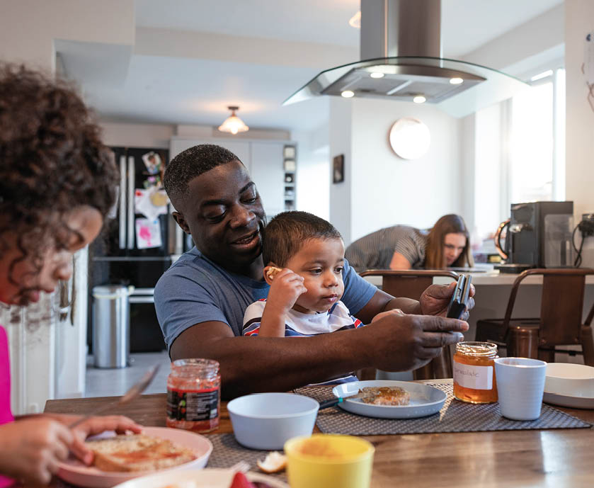 Family having breakfast in the morning together in the North East of England. The father sitting with his young children at the table. The young boy and father are looking at a mobile phone together while the woman is busy in the kitchen behind them. The man is living with a disability after having polio.