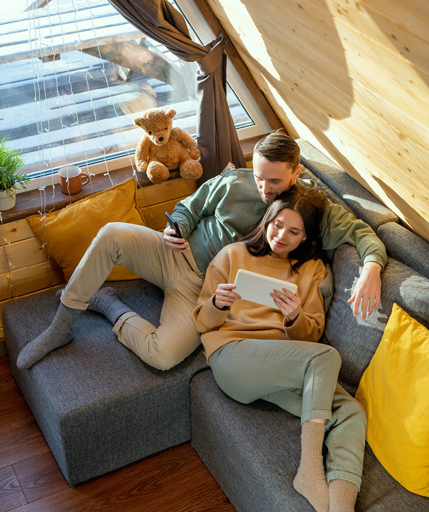 Happy young relaxed husband and wife in casualwear lying on large sofa inside their country house, enjoying rest and watching online movie
