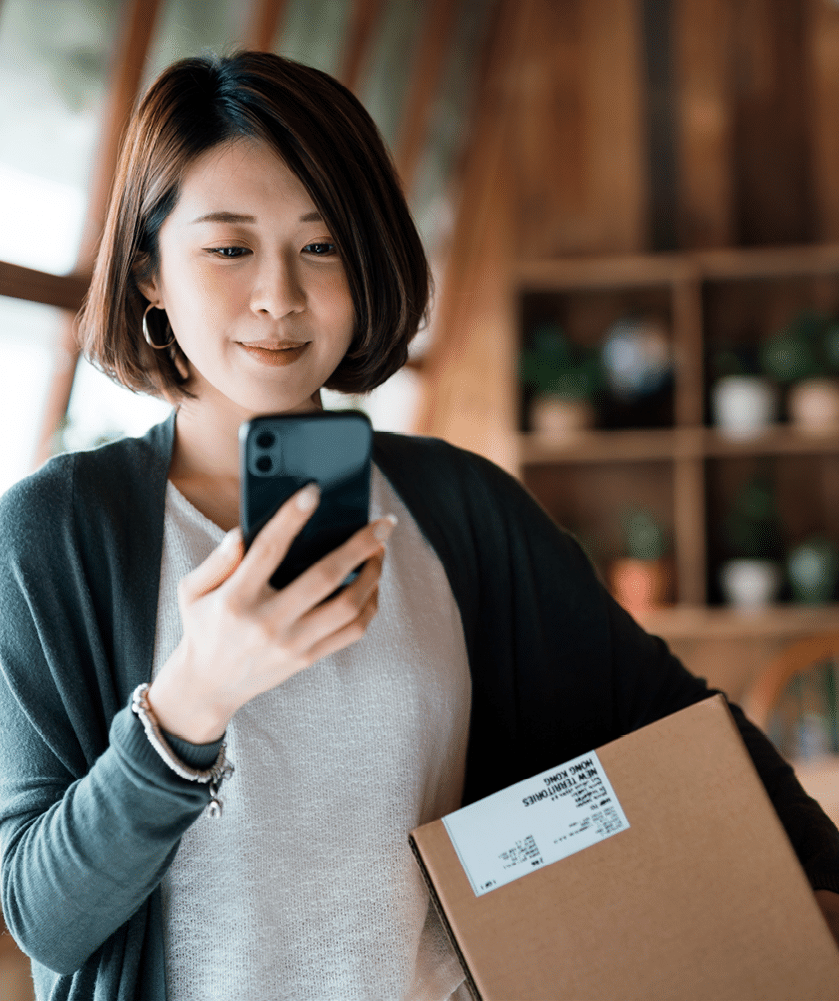 Smiling young Asian woman shopping online with smartphone on hand, receiving a delivered parcel by home delivery service. Online shopping, online banking. Enjoyable customer shopping experience