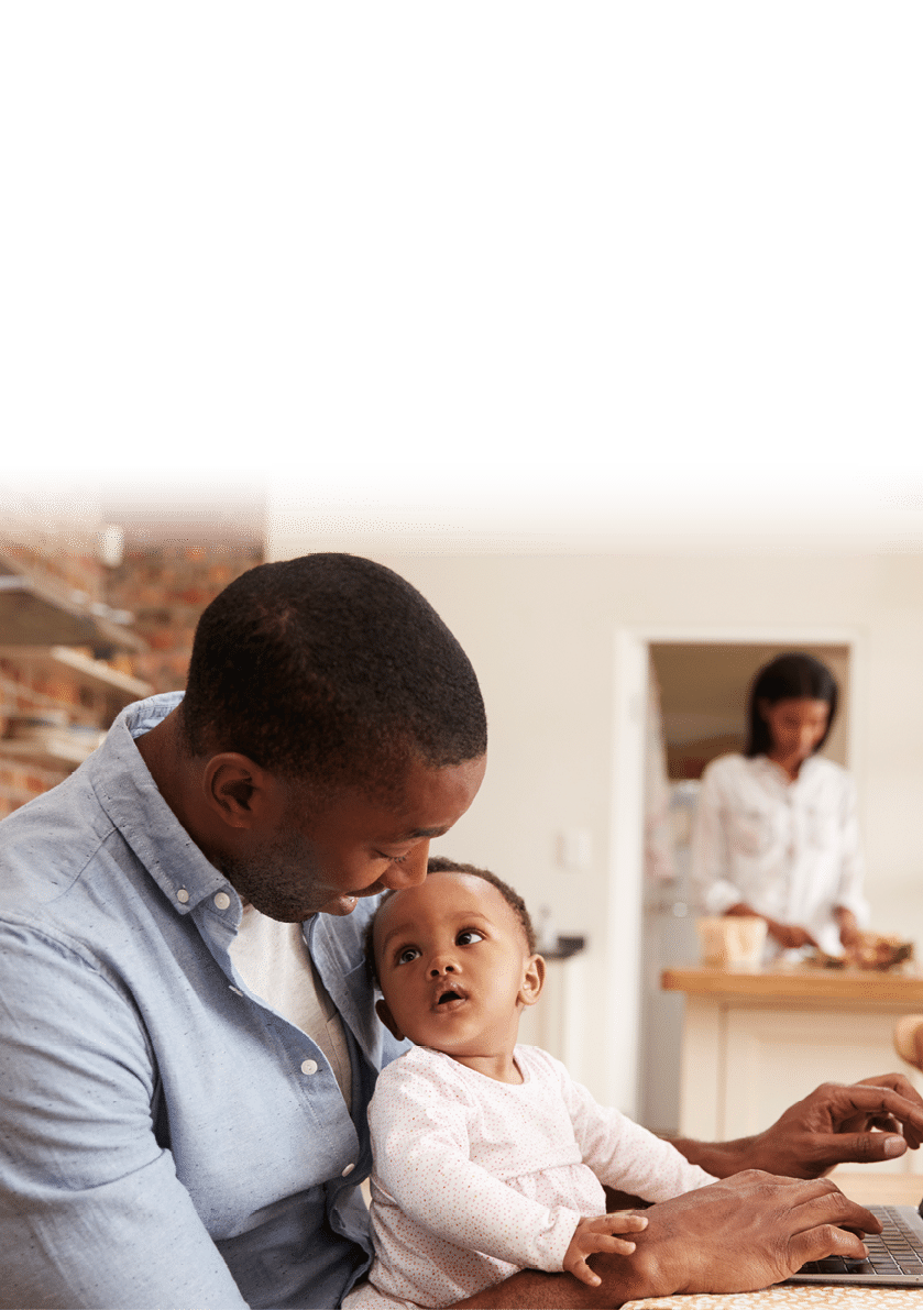 Father And Baby Daughter Use Laptop As Mother Prepares Meal